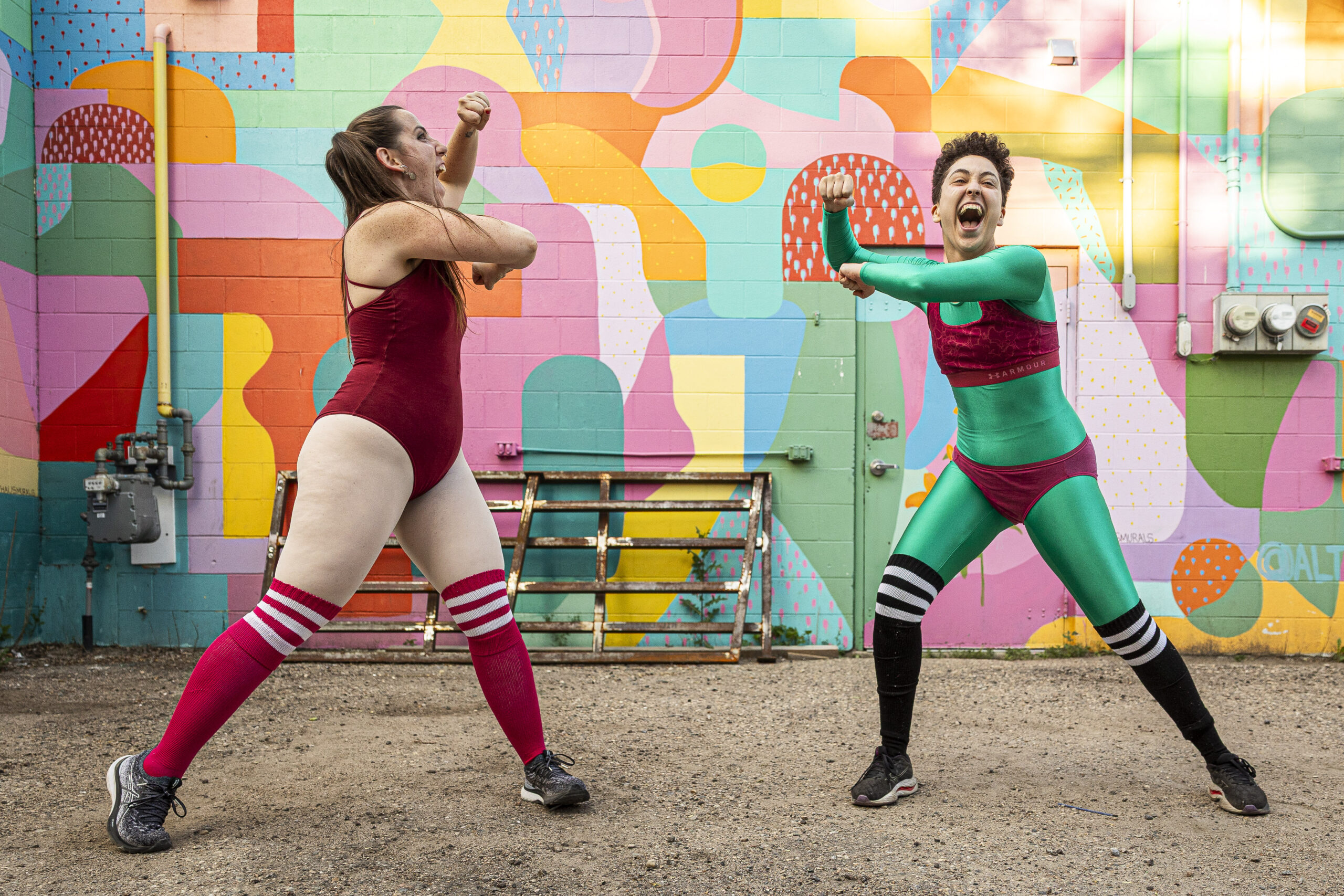 Two dancers in colourful outfits perform in an alley in front of a colourful wall.